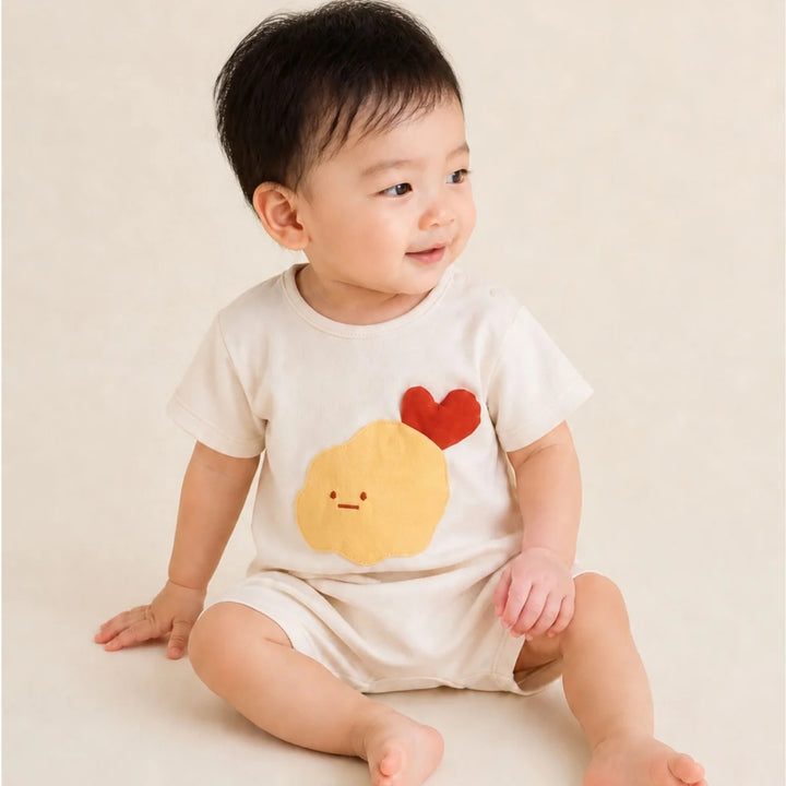 Baby wearing the cream tempura romper with a yellow tempura character and red heart detail, sitting against a soft neutral background.