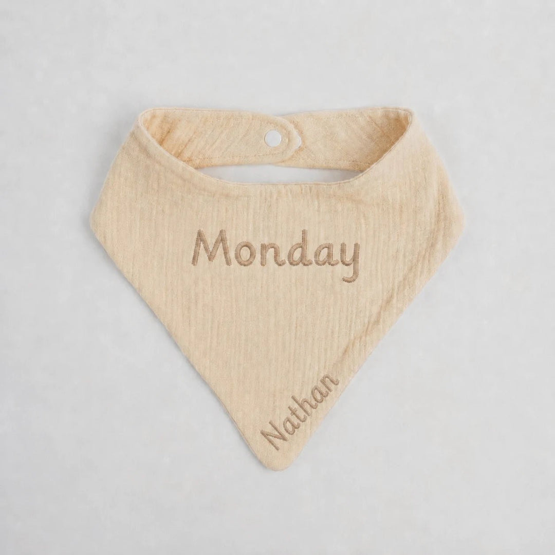 Beige baby bib with 'Monday' and 'Nathan' embroidered on a light gray background