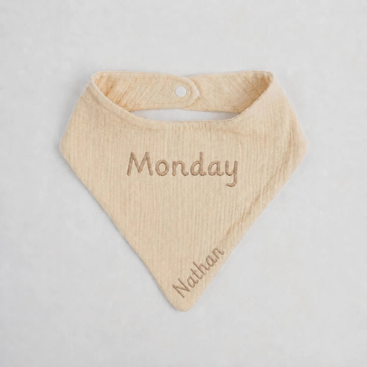 Beige baby bib with 'Monday' and 'Nathan' embroidered on a light gray background