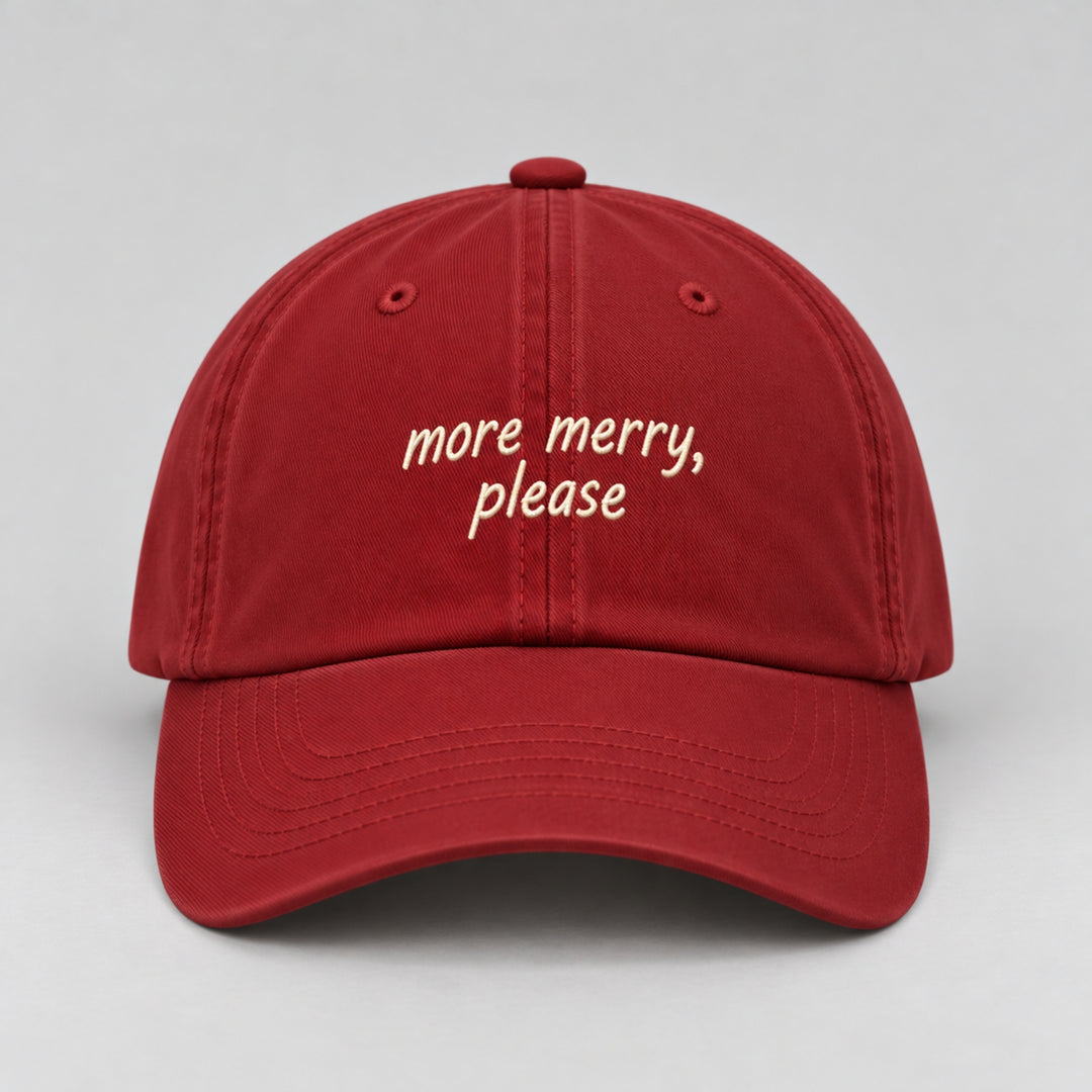 Red Christmas cap embroidered with “more merry, please” in cream thread, featuring a curved brim and minimalist festive design.