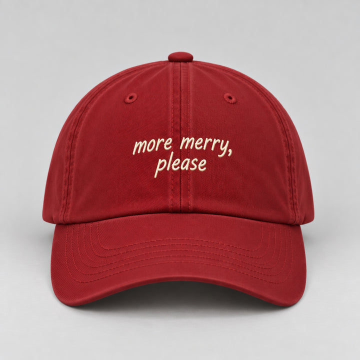 Red Christmas cap embroidered with “more merry, please” in cream thread, featuring a curved brim and minimalist festive design.