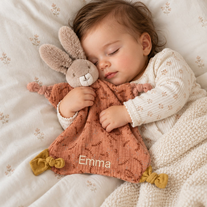 Sleeping baby cuddling a personalised Moulin Roty bunny comforter with terracotta floral fabric and the name Emma embroidered near the bottom.