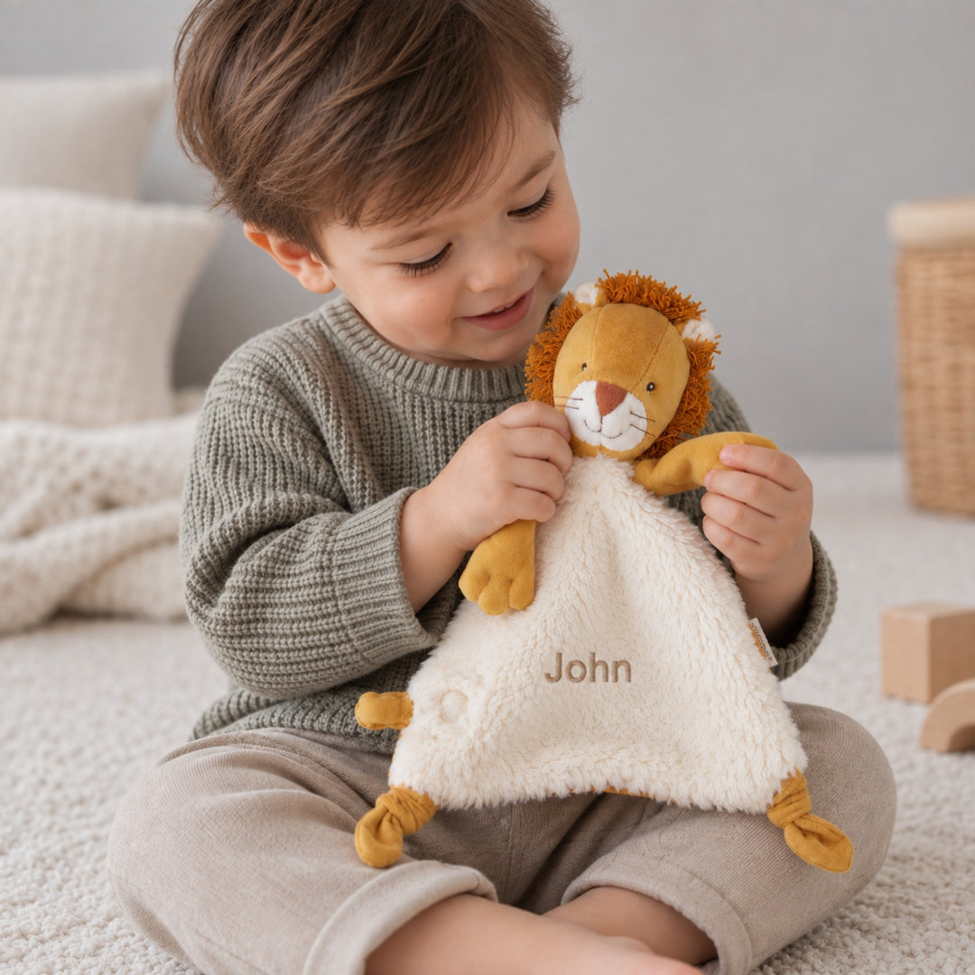 Young boy holding a personalised Moulin Roty Mamba Lion comforter embroidered with the name “John” while sitting in a soft neutral nursery.