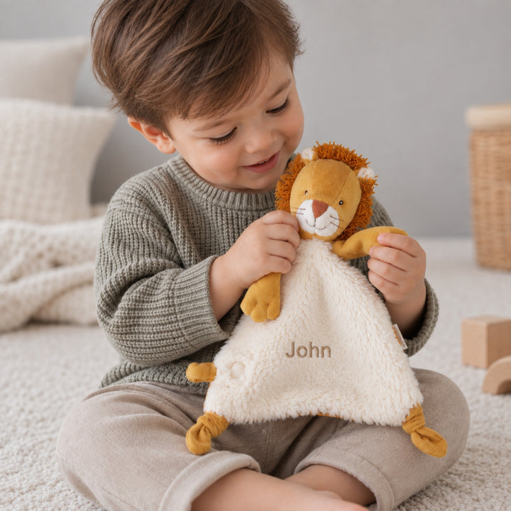 Young boy holding a personalised Moulin Roty Mamba Lion comforter embroidered with the name “John” while sitting in a soft neutral nursery.