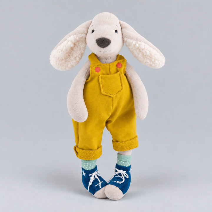Cream plush dog with floppy ears, wearing mustard-yellow overalls, mint-green socks and blue lace-up shoes against a light grey studio background.
