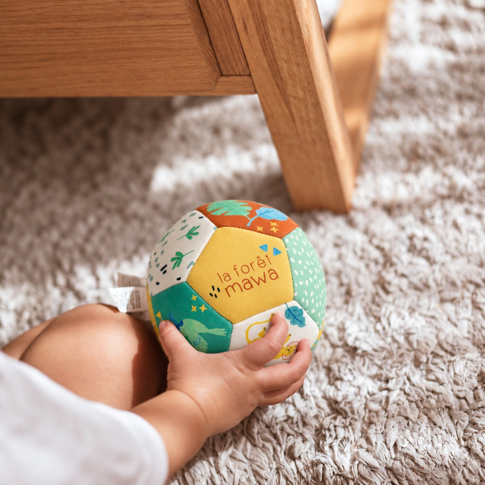 Baby holding the Moulin Roty La Forêt Mawa soft ball while sitting on a textured rug beside wooden furniture.