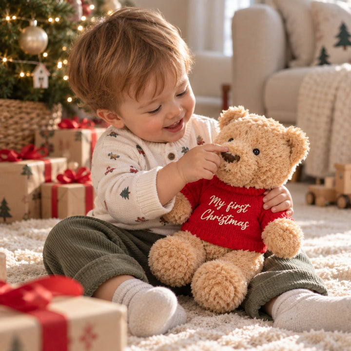 Baby boy sitting by a Christmas tree and playing with a teddy bear wearing a red knitted “My First Christmas” jumper, surrounded by festive presents in a cosy holiday setting.