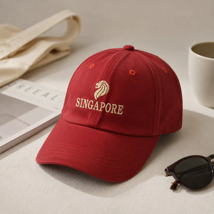 A red baseball cap with beige embroidered lion logo and 'SINGAPORE' text, styled on a tabletop with a book, tote bag, sunglasses, and mug nearby.
