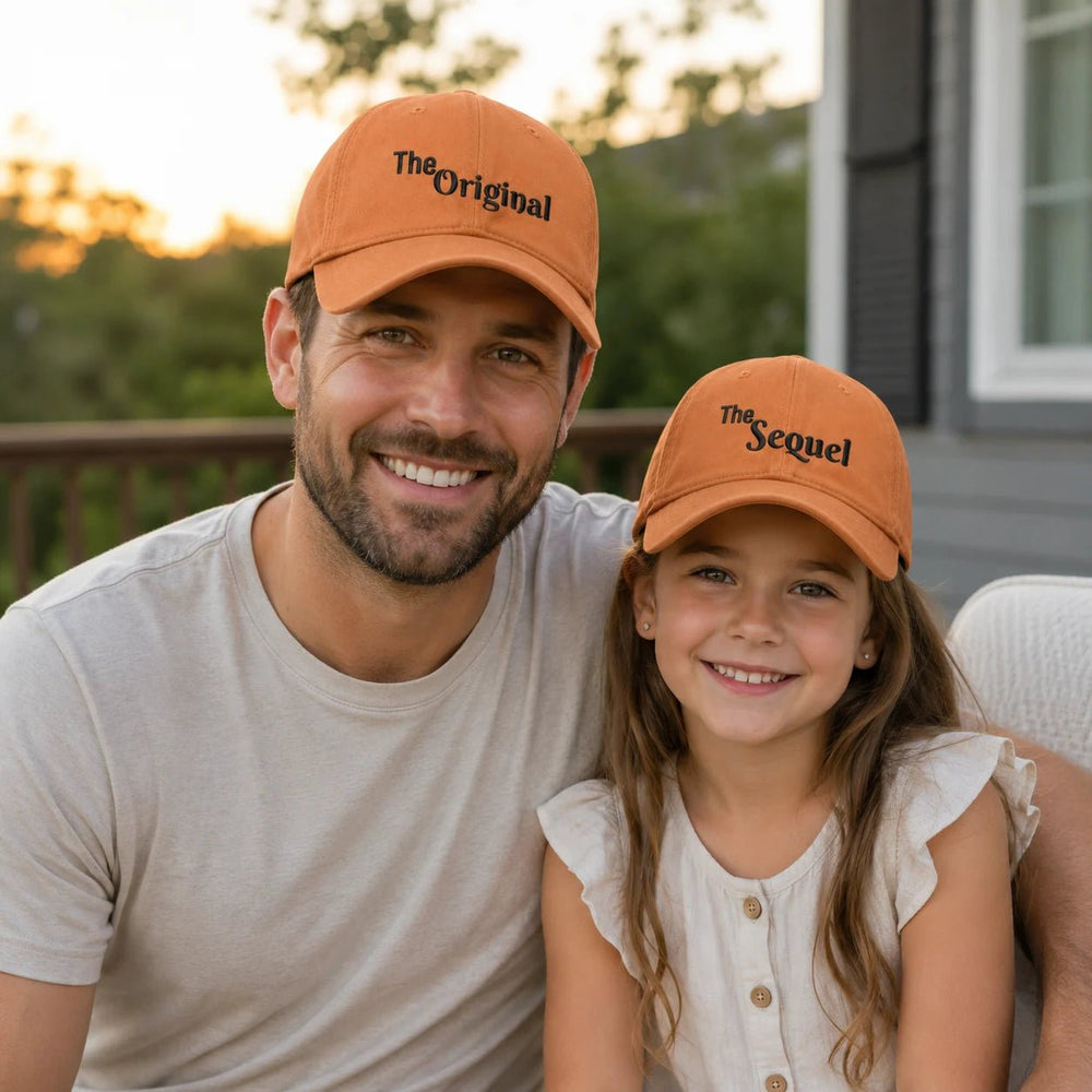 Man and young girl wearing matching orange baseball caps with text, sitting outdoors.