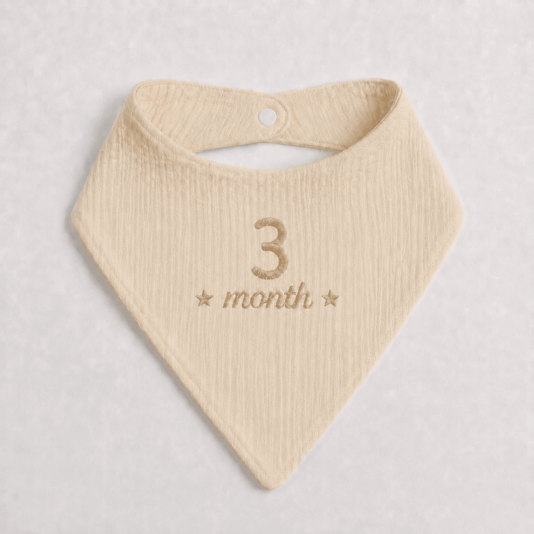 Beige baby bib with '3 month' embroidery on a light gray background