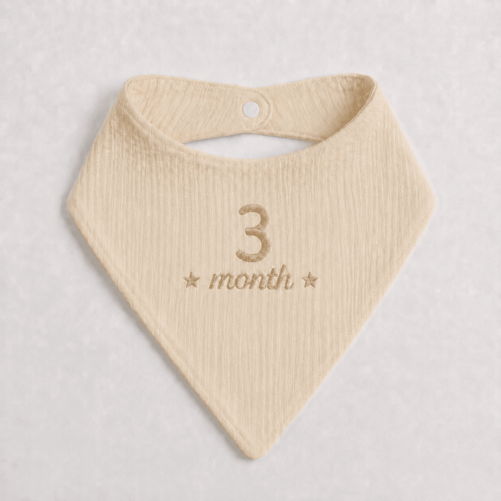 Beige baby bib with '3 month' embroidery on a light gray background