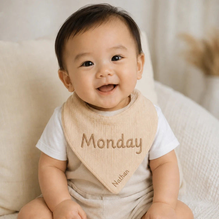 Baby wearing a bib with 'Monday' and 'Nathan' text, sitting on a soft surface.