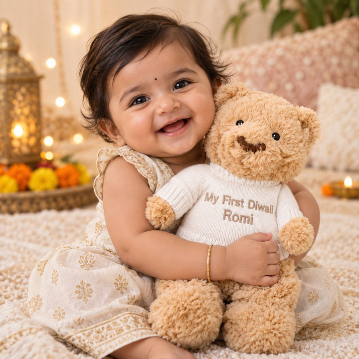 Smiling baby girl cuddling a personalised teddy bear embroidered with “My First Diwali Romi” on a cream knitted jumper in a warm festive Diwali setting.
