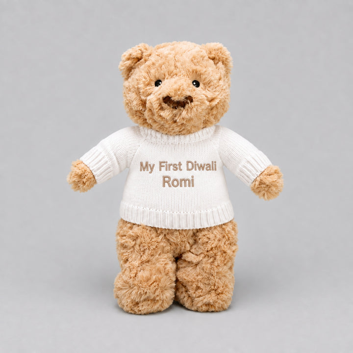 Personalised teddy bear wearing a cream knitted jumper embroidered with “My First Diwali Romi,” photographed on a light grey studio background.