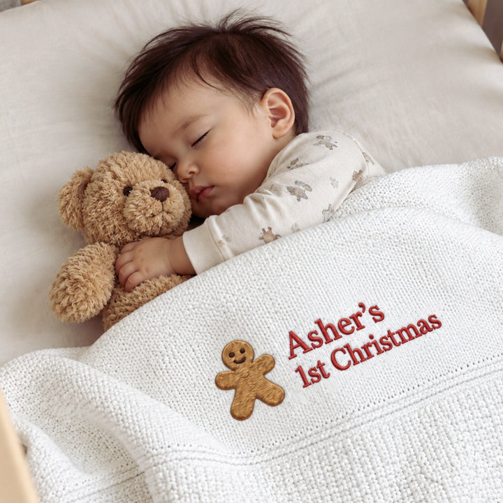 Sleeping baby boy cuddling a soft teddy bear in a cot, covered with a white personalised knitted blanket embroidered “Asher’s 1st Christmas” with a gingerbread motif.