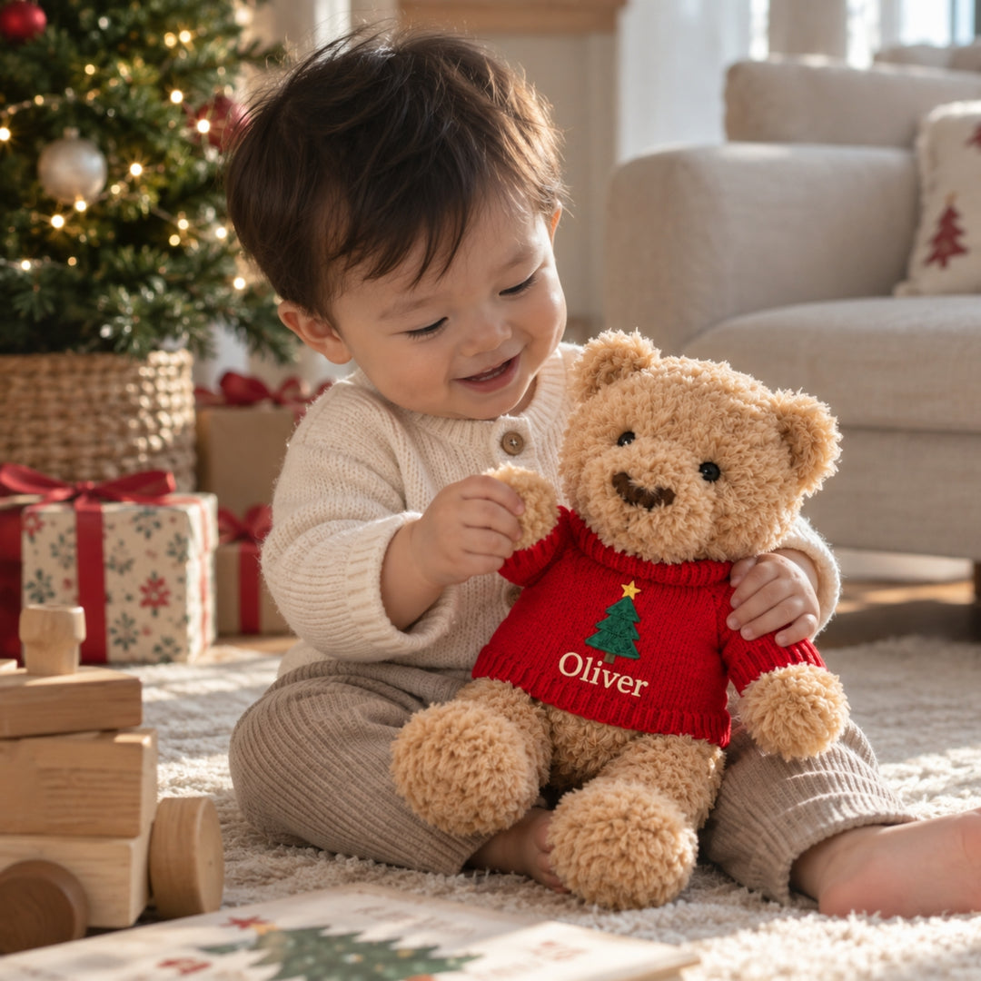 Baby boy playing with a personalised teddy bear wearing a red Christmas jumper embroidered with a Christmas tree and the name “Oliver”, in a cosy festive setting with a Christmas tree and presents.