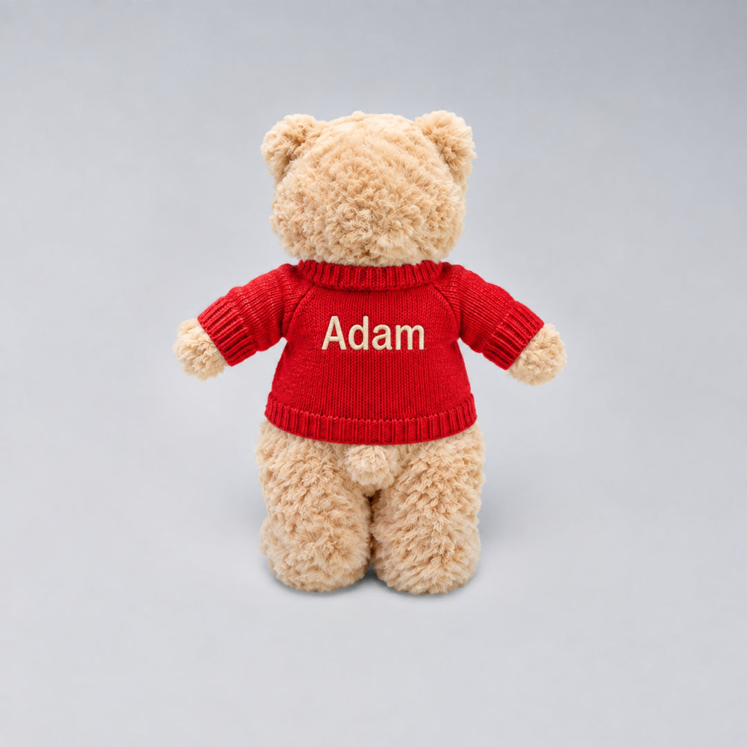 Back view of a personalised Christmas teddy bear wearing a red knitted jumper embroidered with the name “Adam” in cream, photographed against a light grey studio background.
