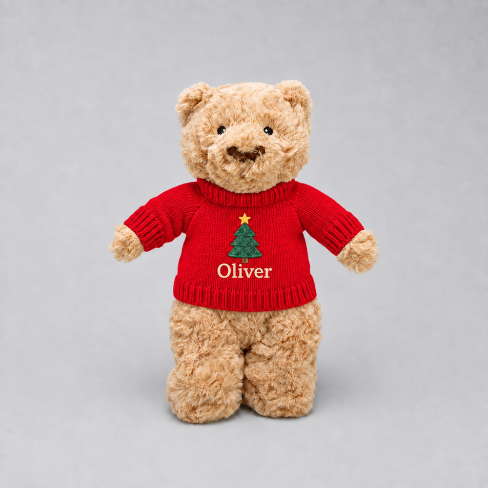 Personalised teddy bear wearing a red knitted Christmas jumper embroidered with a festive Christmas tree and the name “Oliver”, photographed against a light grey studio background.