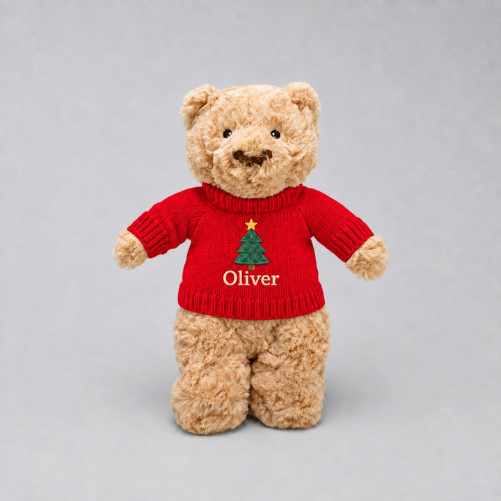 Personalised teddy bear wearing a red knitted Christmas jumper embroidered with a festive Christmas tree and the name “Oliver”, photographed against a light grey studio background.