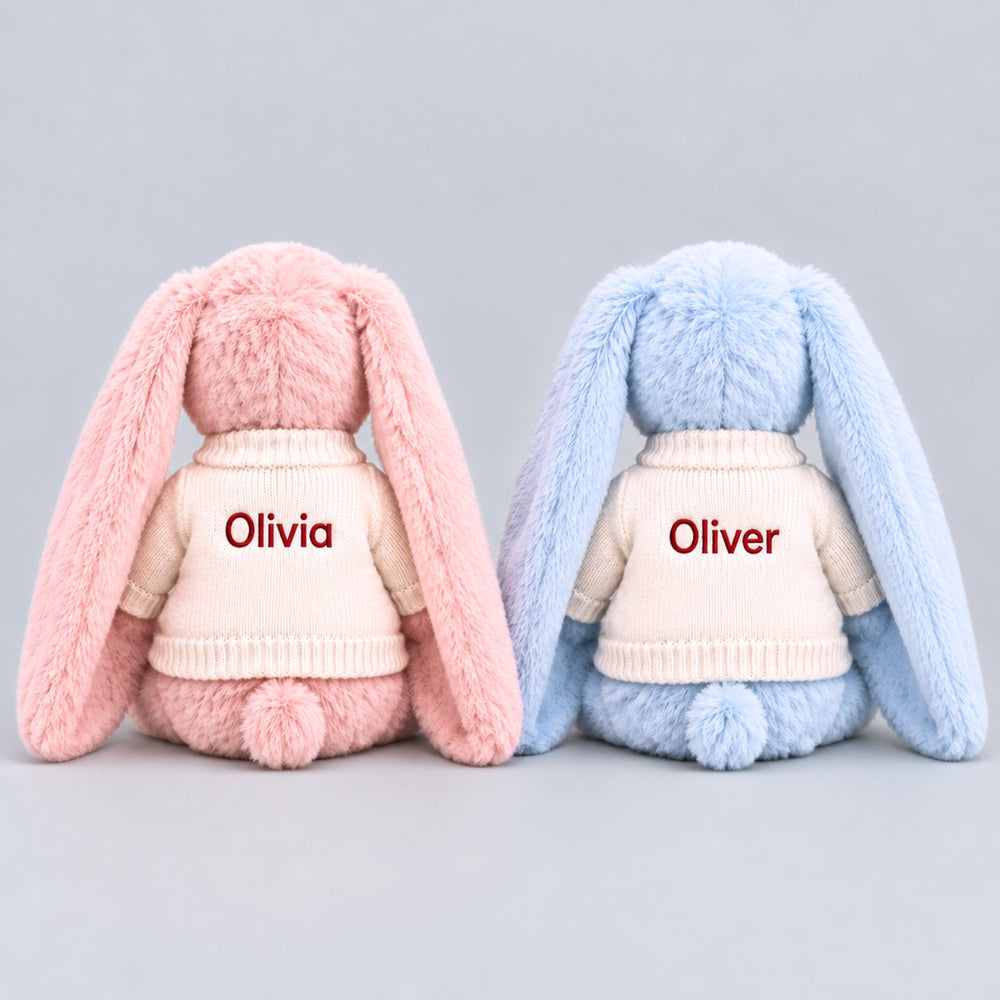 Back view of two personalised plush bunnies, one pink and one blue, wearing cream knitted jumpers embroidered with “Olivia” and “Oliver” in red thread, featuring long floppy ears and small fluffy tails against a light grey studio background.