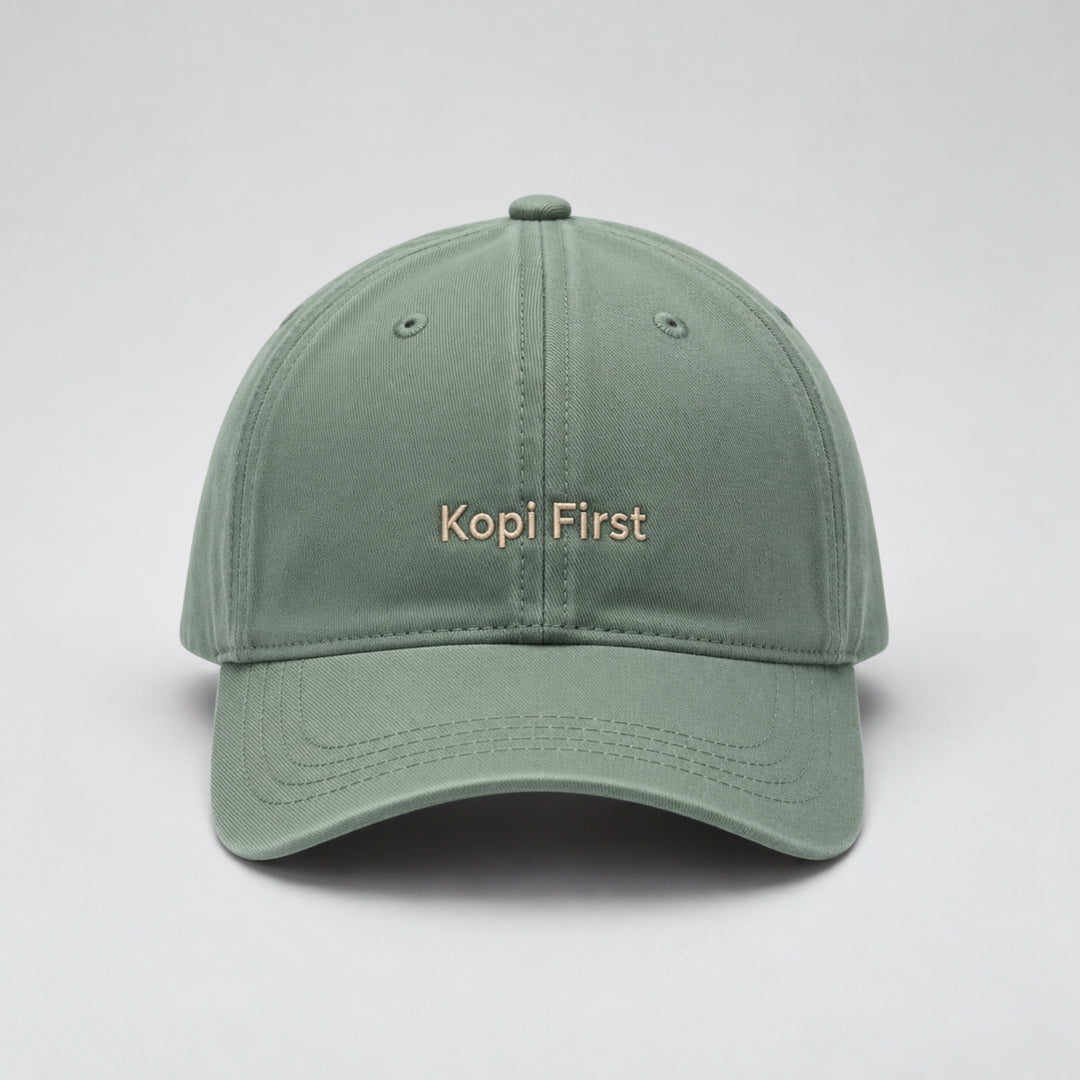 Sage green baseball cap embroidered with “Kopi First” in beige block lettering, featuring a curved brim and classic six-panel design against a light grey studio background.
