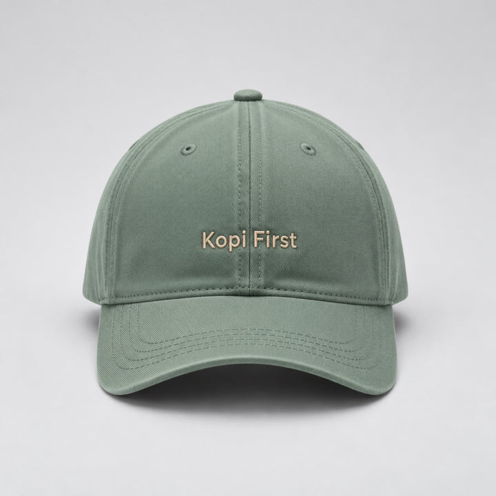 Sage green baseball cap embroidered with “Kopi First” in beige block lettering, featuring a curved brim and classic six-panel design against a light grey studio background.