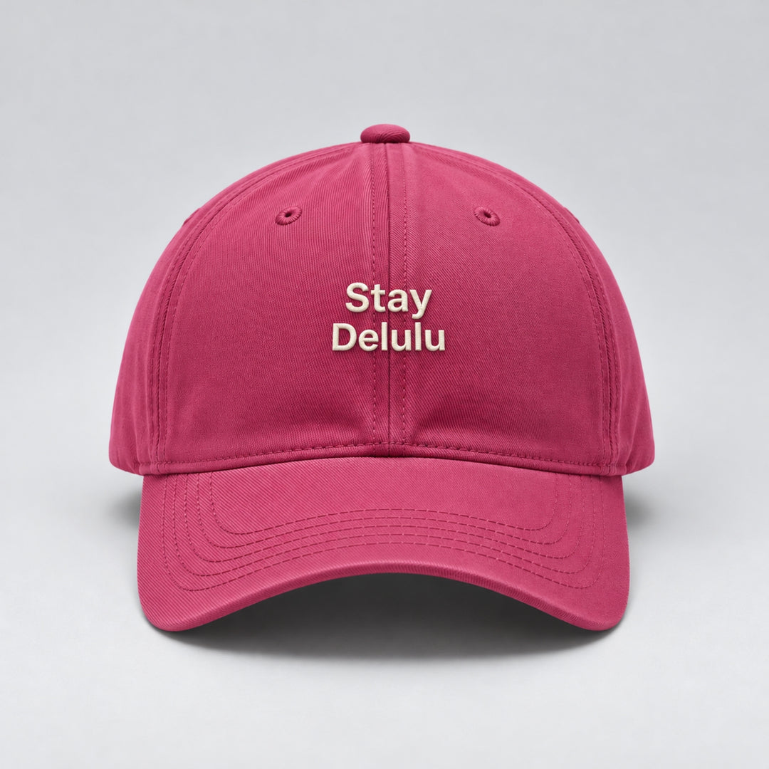 Dark pink baseball cap featuring “Stay Delulu” embroidered in cream block lettering across two lines, with a curved brim and classic six-panel design.