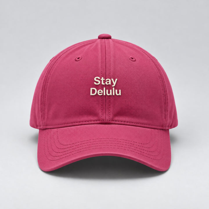 Dark pink baseball cap featuring “Stay Delulu” embroidered in cream block lettering across two lines, with a curved brim and classic six-panel design.
