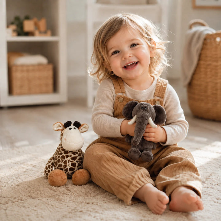 A smiling toddler sits on a light rug in a bright nursery, holding a dark gray elephant plush toy and sitting next to a giraffe plush toy. The child is wearing tan corduroy overalls over a cream long-sleeve shirt.