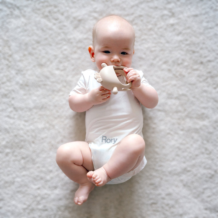 Baby in White babygrow and biting beige chick teether
