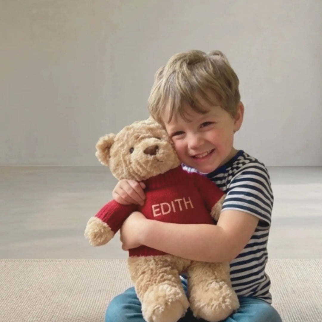 Child holding a teddy bear with a red shirt labeled 'Edith' on a neutral background