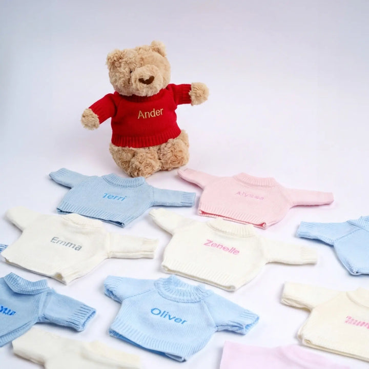 Collection of small sweaters with embroidered names around a teddy bear wearing a red sweater.