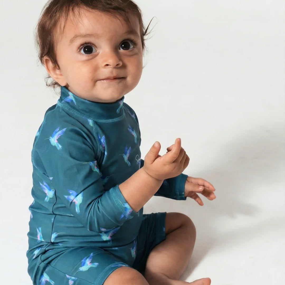 Baby sitting and wearing the teal hummingbird-print long-sleeve swimsuit with high neckline and short-leg design.