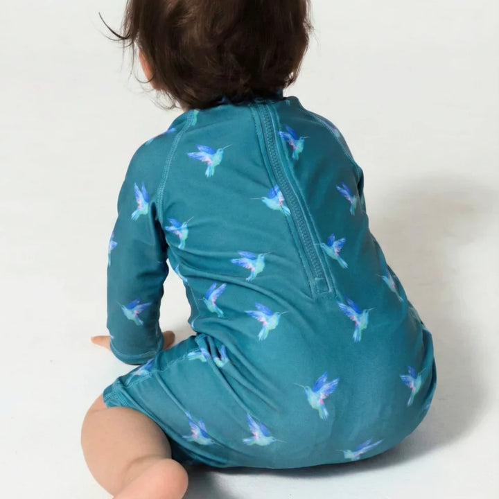 Baby wearing the teal hummingbird-print swimsuit from the back, showing the rear zip fastening and all-over bird pattern.