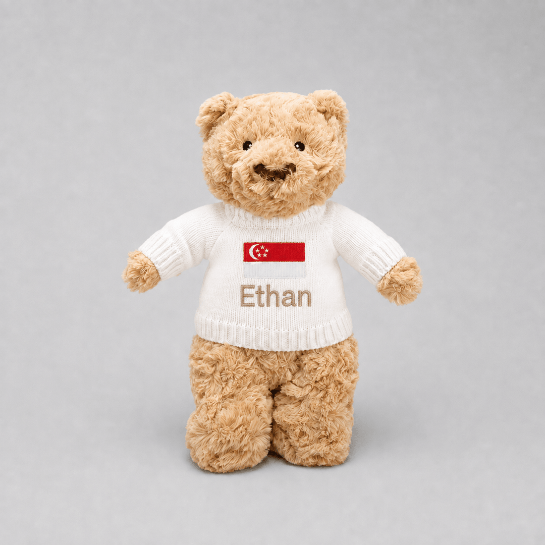 A plush teddy bear in light brown fur wearing a white knit sweater with a red Singapore flag patch and the name 'Ethan' embroidered on the front, photographed against a neutral gray background.