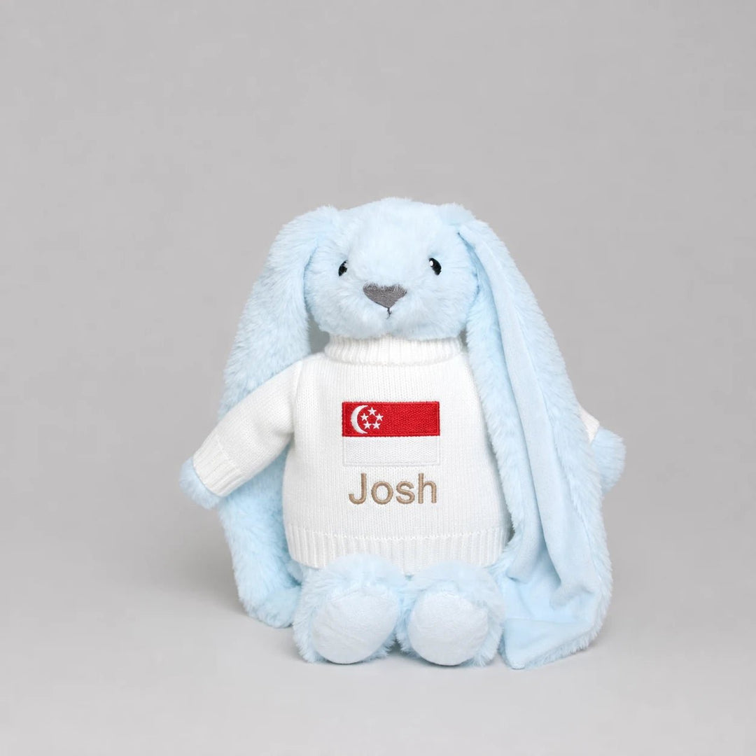 A light blue plush bunny toy wearing a white knit sweater with a Singapore flag patch and the name 'Josh' embroidered on the front, photographed against a neutral gray background.