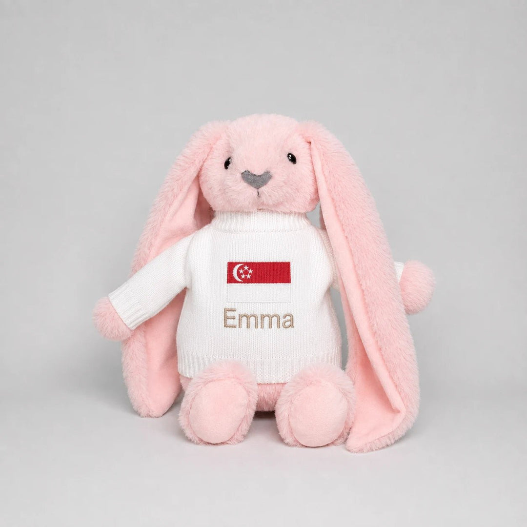 A pink plush bunny toy wearing a white knit sweater with a Singapore flag patch and the name 'Emma' embroidered on the front, photographed against a light gray studio background.