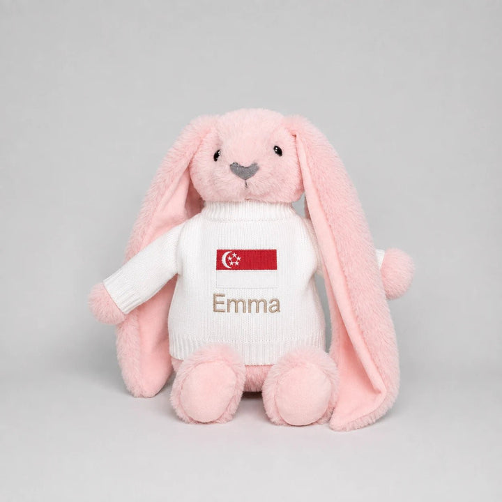 A pink plush bunny toy wearing a white knit sweater with a Singapore flag patch and the name 'Emma' embroidered on the front, photographed against a light gray studio background.