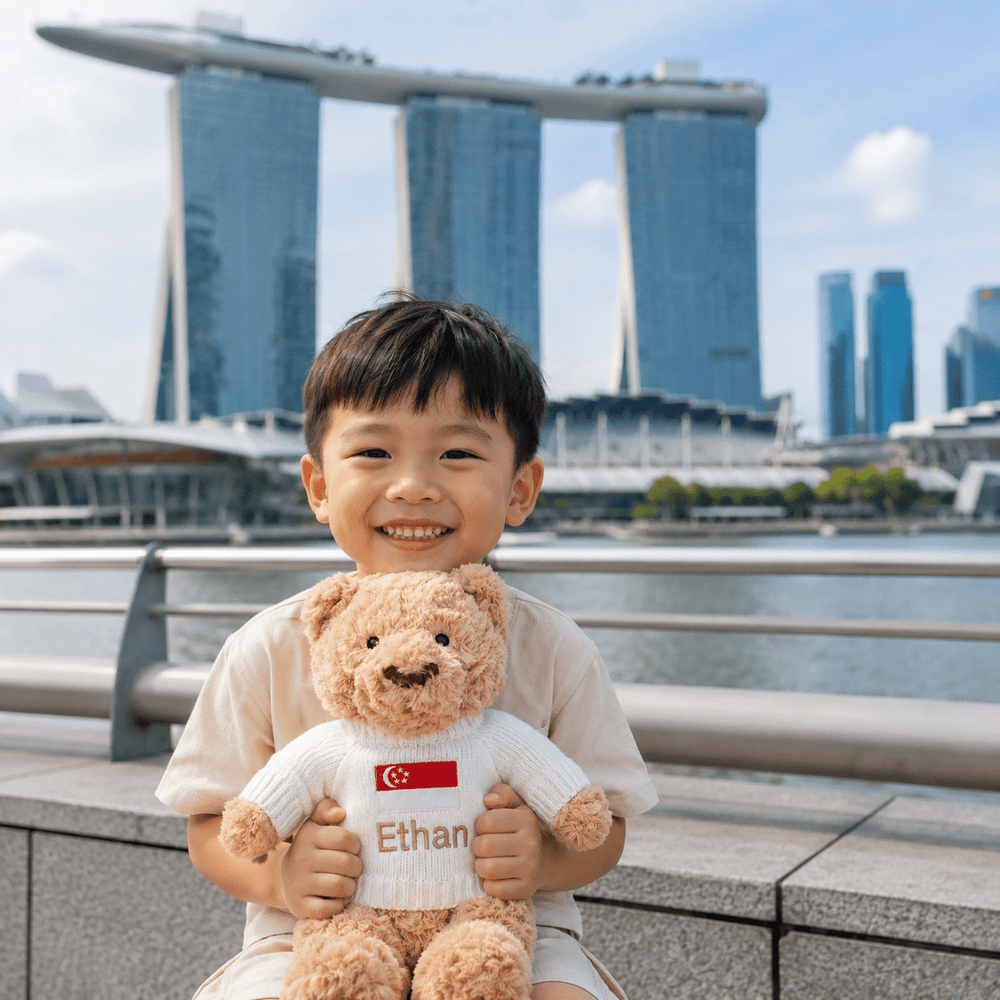 A smiling young boy stands outdoors in front of a waterfront city skyline, holding a teddy bear. The teddy bear wears a white sweater with a Singapore flag patch and the name 'Ethan' printed on it.