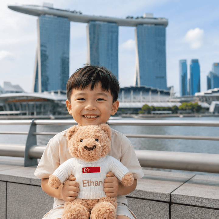 A smiling young boy stands outdoors in front of a waterfront city skyline, holding a teddy bear. The teddy bear wears a white sweater with a Singapore flag patch and the name 'Ethan' printed on it.