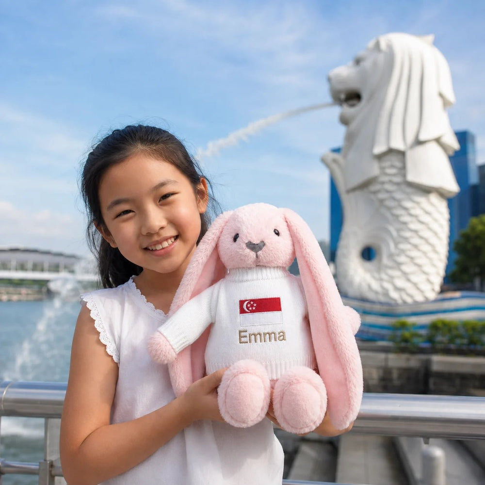 A young girl smiling while holding a pink plush bunny sweater in front of the Merlion statue in Singapore. The bunny is wearing a white knit top with a Singapore flag patch and the name 'Emma' embroidered on the front.