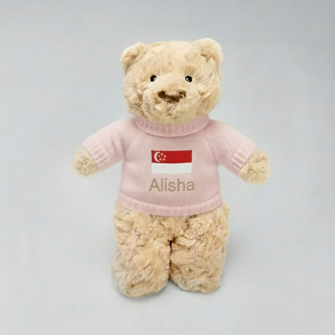 Beige teddy bear wearing a pink sweater with 'Alisha' and a Singapore flag on a light gray background