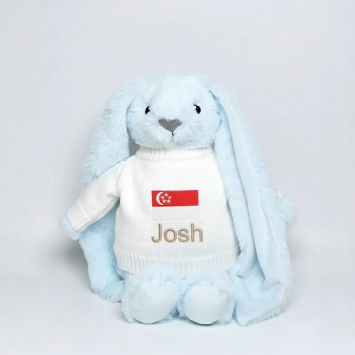 Blue plush bunny with a white sweater displaying a Singapore flag and the name 'Josh' on a light gray background