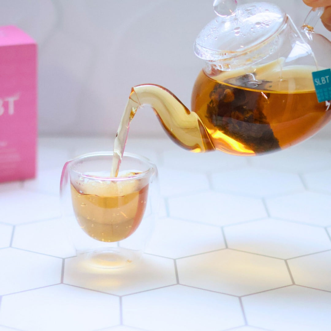Golden nursing tea being poured from a glass teapot into a clear cup, with an SLBT tea bag steeping inside.