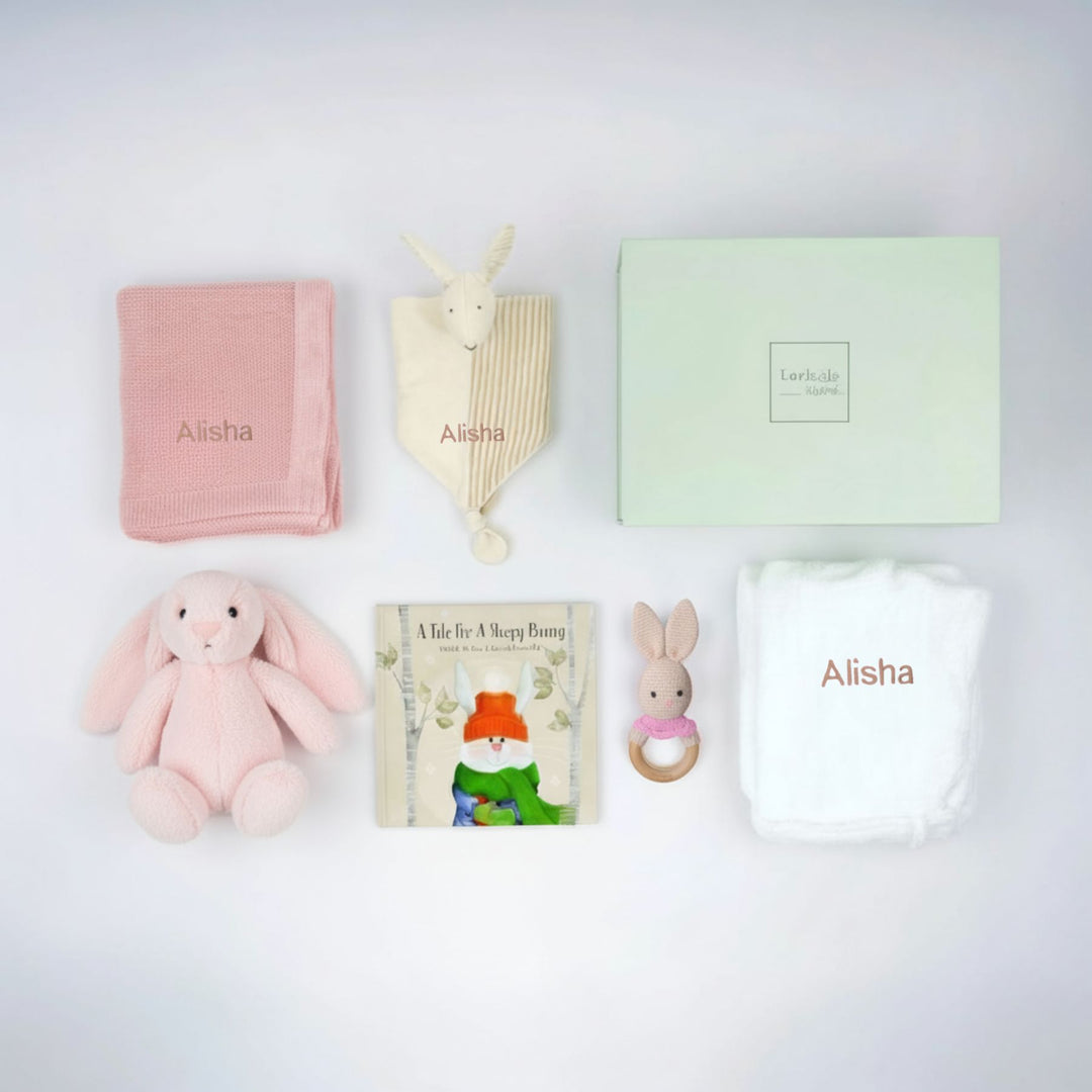 Collection of baby items including a pink blanket, plush toys, and a book on a light gray background.