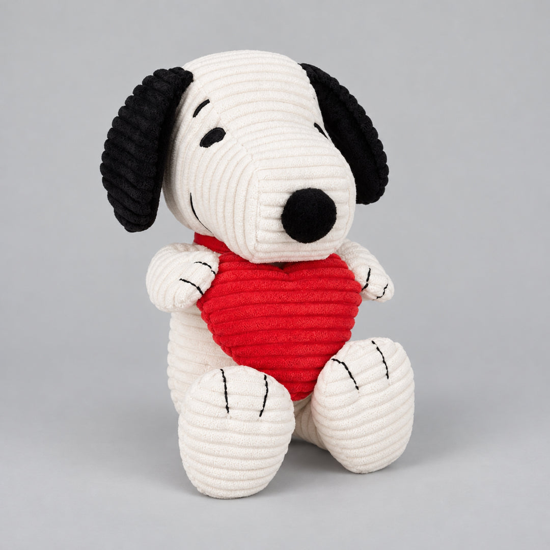 White Snoopy plush toy with black floppy ears, holding a bright red textured heart while sitting against a light grey background.