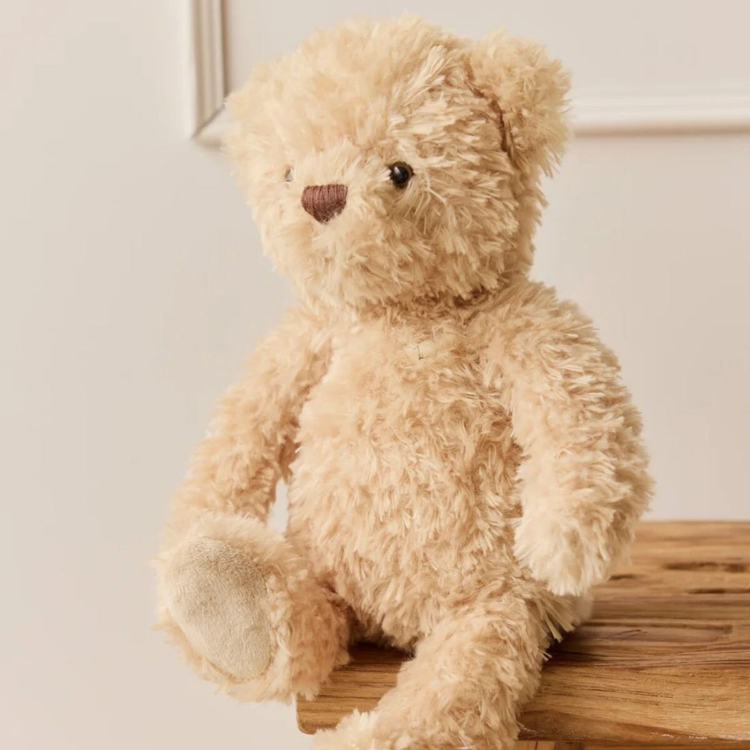 Soft cream plush Arthur teddy bear with fluffy textured fur, dark eyes and a brown embroidered nose, sitting on a wooden bench in a cosy nursery setting.