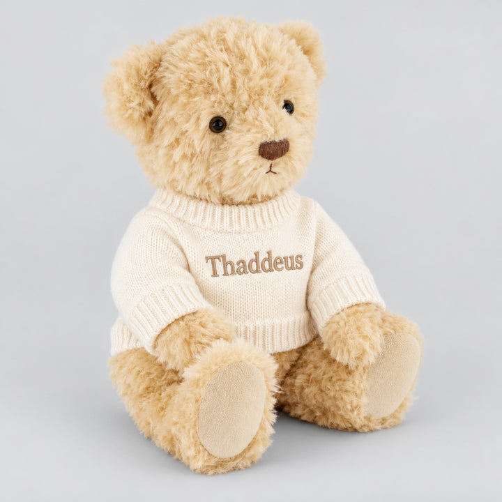 Personalised cream Arthur teddy bear wearing a white knitted jumper embroidered with the name Thaddeus in beige thread, seated against a light grey studio background.