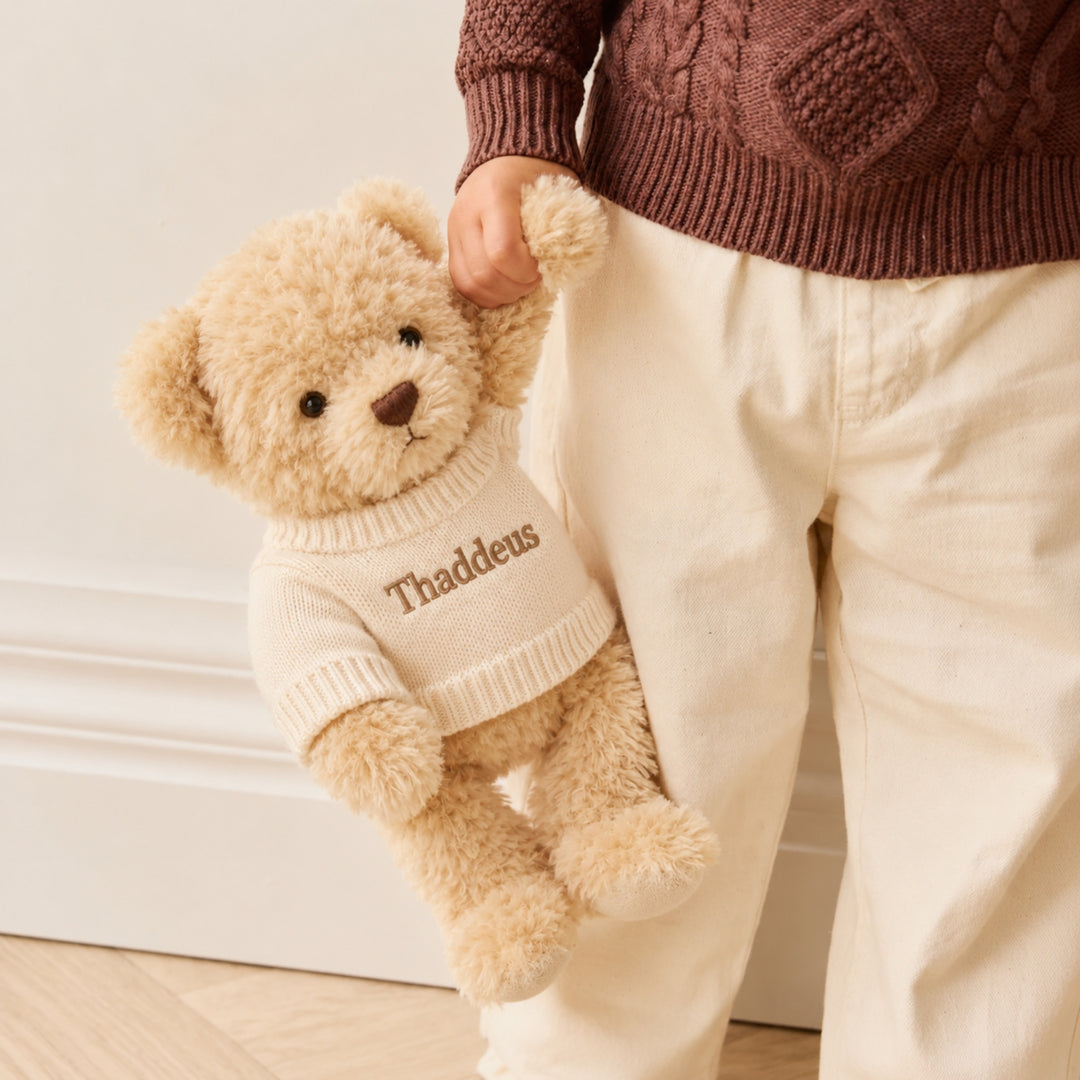 Young child holding a fluffy cream Arthur teddy bear dressed in a personalised white knitted jumper with the name Thaddeus, wearing a brown sweater and cream trousers.