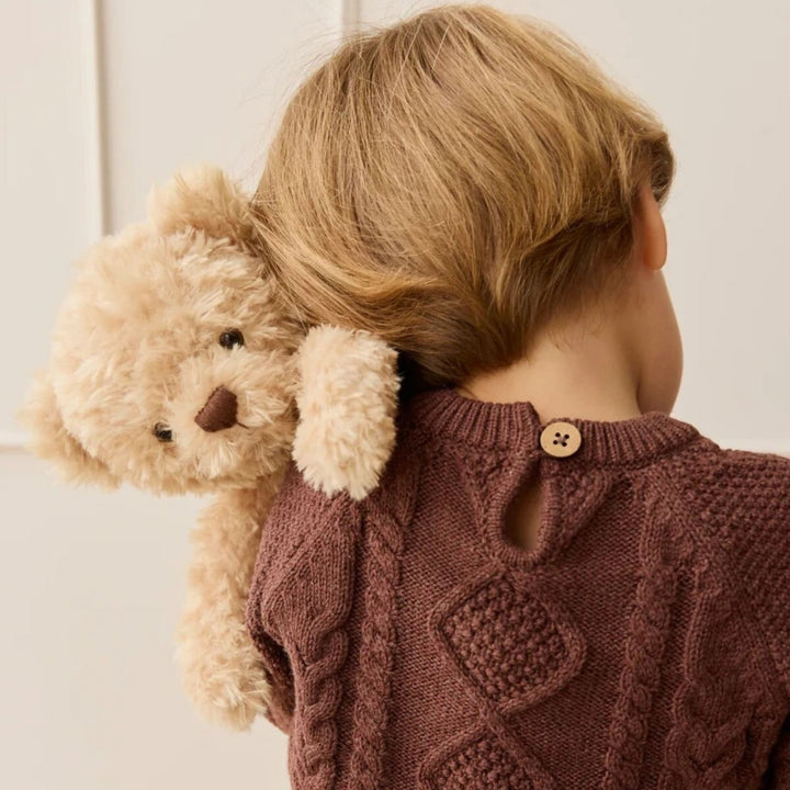 Young child carrying a soft cream Arthur teddy bear over their shoulder, dressed in a brown cable-knit jumper in a warm, neutral home setting.